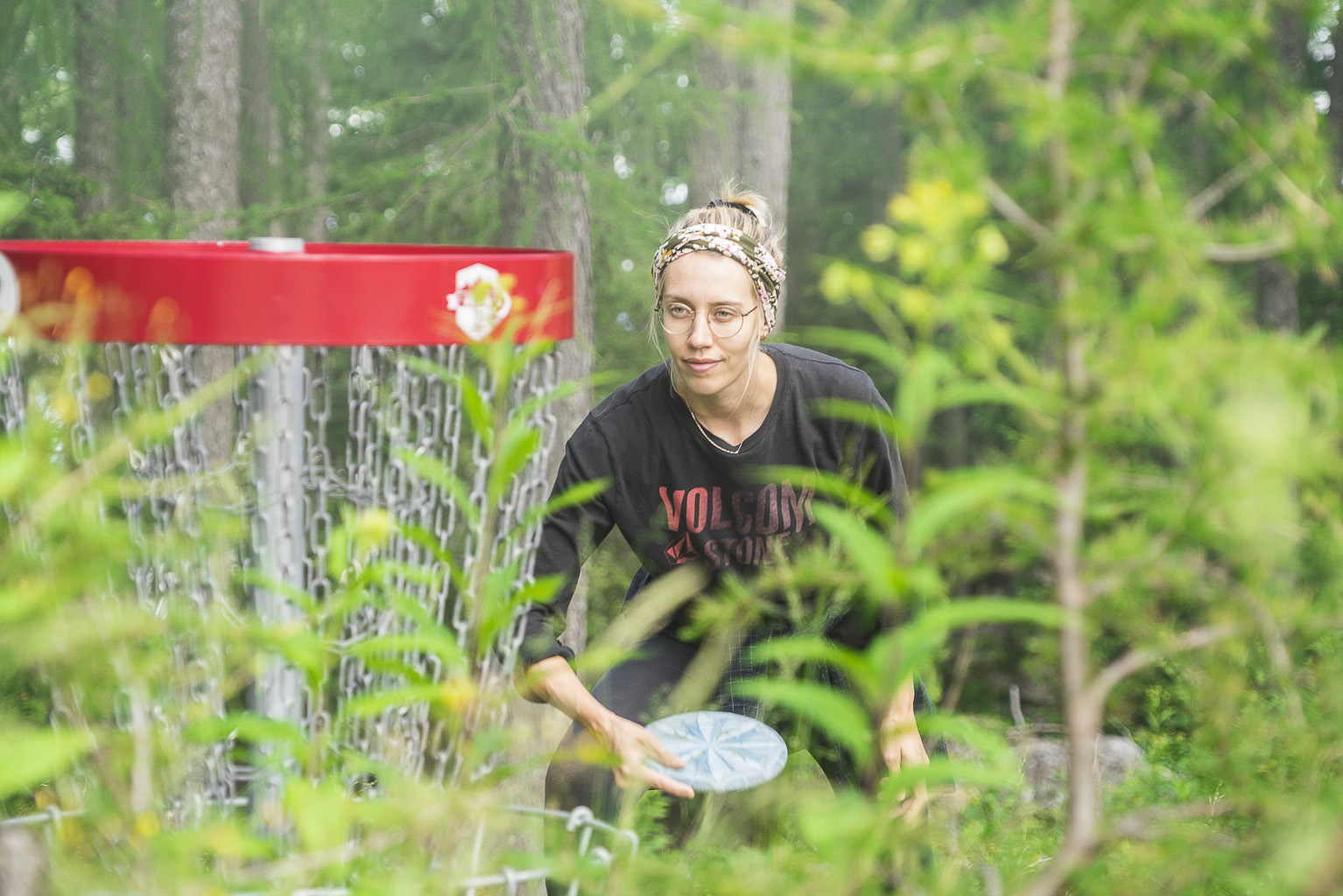 Eine junge Frau spielt Discgolf. Sie steht wenige Meter vor dem Korb, hält eine blaue Disc in der Hand und zielt auf den Korb.